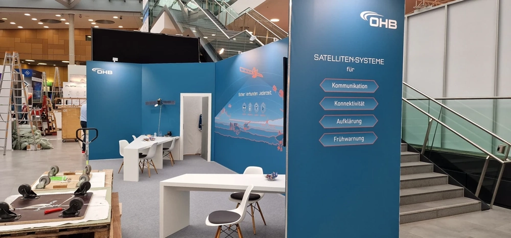 Blauer Messestand im Aufbau mit Trennwänden, Tischen, Stühlen und Modulen – Messestandbau