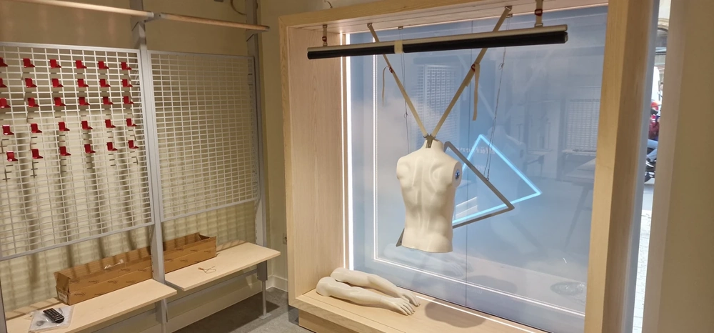Schaufenster mit hellem Holzrahmen, hängender Torso-Mannequinfigur und liegenden Beinelementen auf Podest, Showroom montage