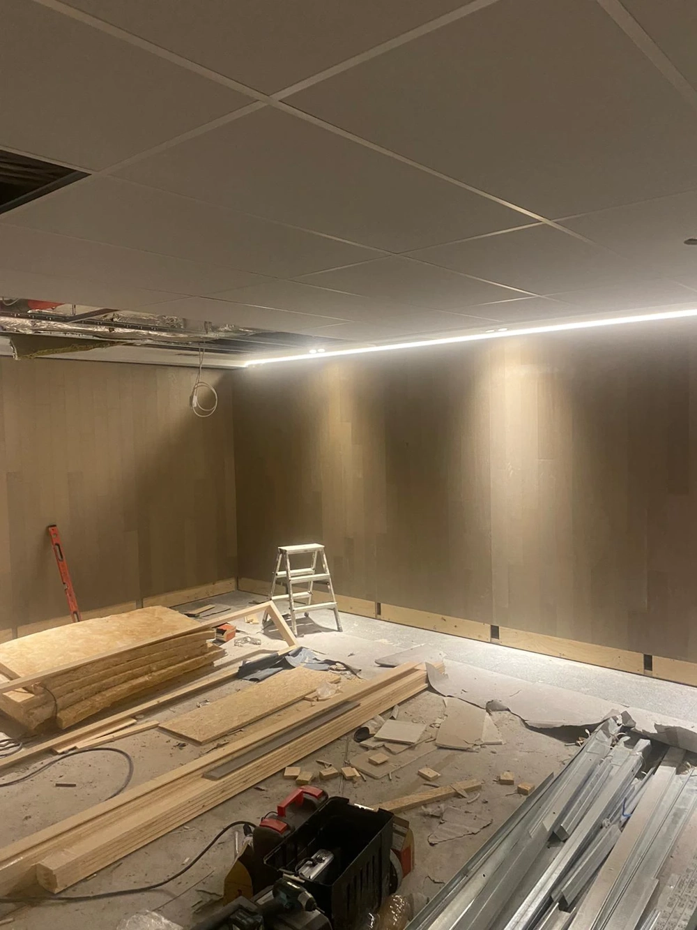 Unfertiger Innenraum mit installierter Rasterdecke, Holzplatten an Wand, Leiter und Baumaterial auf Boden – Rasterdecke