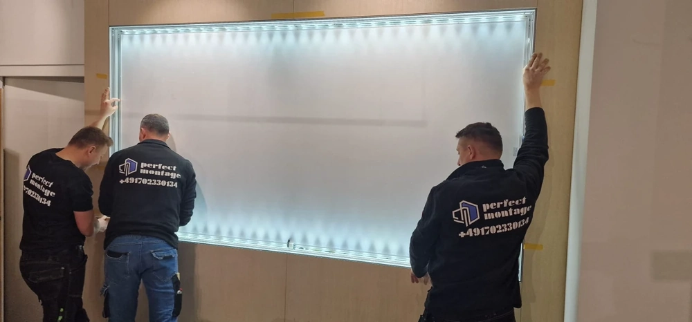 Drei Monteure montieren eine beleuchtete Wandplatte auf Holzunterkonstruktion beim Messestandbau