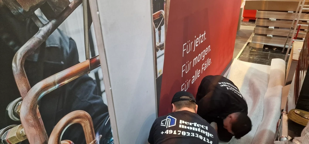 Zwei Monteure verlegen Teppichboden an einem Messestand hinter rotem Werbebanner