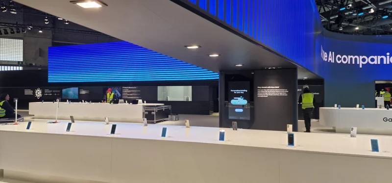 Messestandbau einer modernen Messeinstallation mit langer weißer Theke, Display-Geräten und digitaler Leuchtrückwand