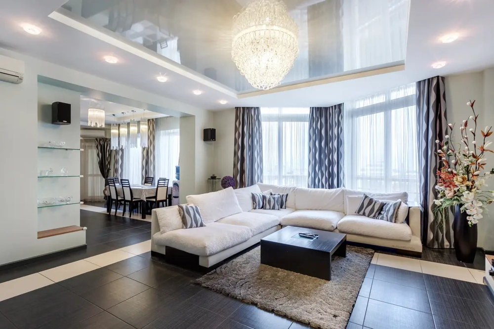 Modernes Wohnzimmer mit weißem Ecksofa, dunklem Couchtisch, Teppich, Kronleuchter, Vorhängen und offenem Essbereich