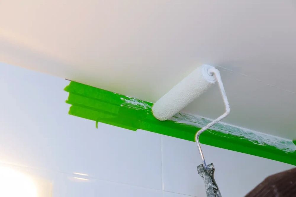 Malerarbeiten: Weißer Farbroller verteilt Farbe an abgeklebte Decke mit grünem Malerkrepp